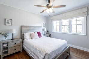 Ein Schlafzimmer mit einem hölzernen Bett, weißen Wänden, einem Deckenventilator, einem Fenster mit weißen Vorhängen und einem Nachttisch mit Lampe und Vase.