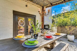 An outdoor dining area with a long table set for a meal, a grill, and a view of a garden. - Saint Elmo Loft | Upscale Retreat • Near Downtown! (Chattanooga)