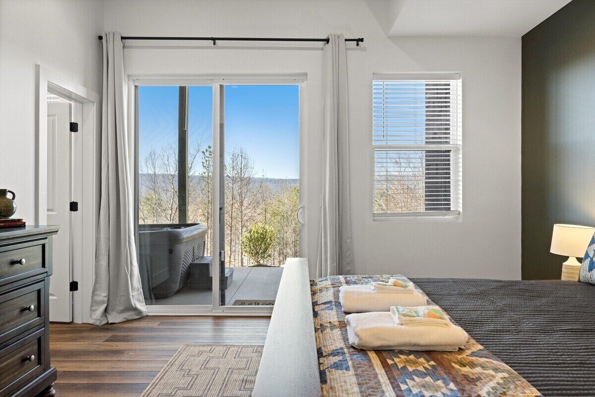 A bedroom with a bed, a nightstand, a lamp, a window with blinds, and a view of the outdoors.