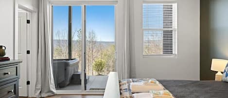 A bedroom with a bed, a nightstand, a lamp, a window with blinds, and a view of the outdoors.