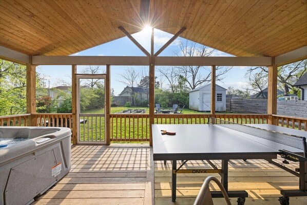 A covered outdoor area with a hot tub, ping pong table, and a view of a backyard.