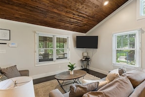 A living room with a wooden ceiling, two windows with blinds, a flat-screen TV, a round coffee table with a plant, and a sofa.