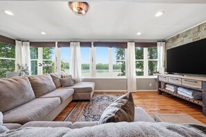 A spacious living room with a large sectional sofa, a flat-screen TV, and a large window with a view of greenery. - Lake front/Hot Tub/Game Room/Dock  Home in Lexington, NC (Lexington)