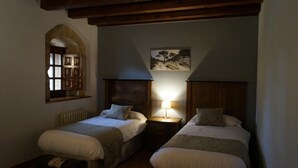 Une chambre avec deux lits, une tête de lit en bois, une petite table avec une lampe et une fenêtre donnant sur des arbres.