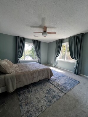 Quarto com cama, ventilador de teto e janelas amplas com cortinas.