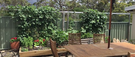 Een terras buiten met een houten tafel en stoelen, een grote parasol en een tuin met verschillende planten en bloemen.