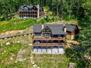A two-story lodge with a stone facade, large windows, and a balcony overlooking a forested area. - Love & Sunsets at Eagles Nest - hot tub, expansive mountain view, outdoor fireplace (Banner Elk)