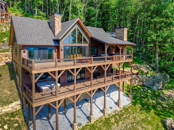 A multi-level wooden cabin with large windows, a deck, and a stone foundation. - Love & Sunsets at Eagles Nest - hot tub, expansive mountain view, outdoor fireplace (Banner Elk)