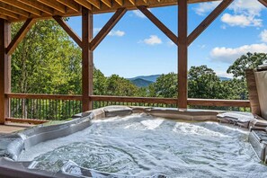 A hot tub with a wooden deck and railing, surrounded by trees and a mountain view. - Love & Sunsets at Eagles Nest - hot tub, expansive mountain view, outdoor fireplace (Banner Elk)
