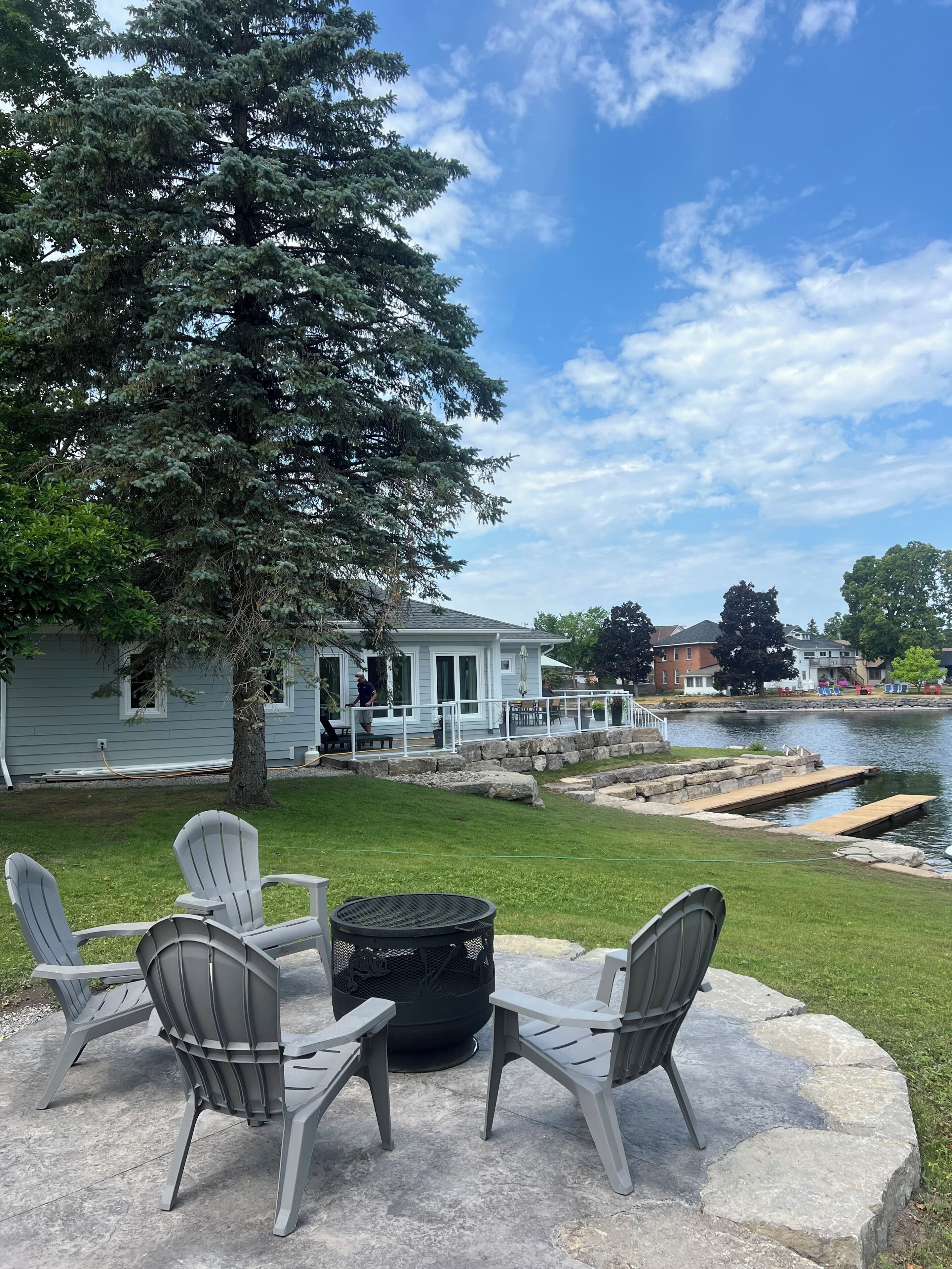 Eine Terrasse mit Adirondack-Stühlen und einem Feuerkorb mit Blick auf einen See, an dem eine Anlegestelle und ein Haus im Hintergrund liegen.