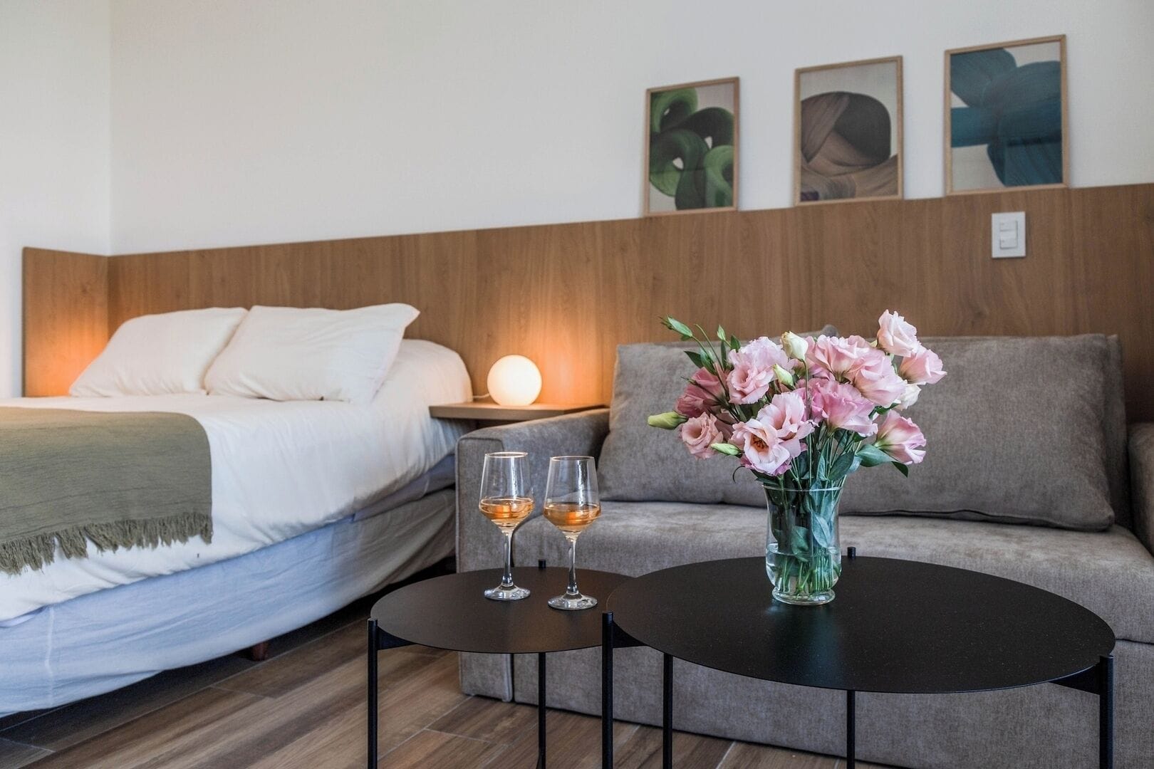Een hotelkamer met een bed, een bank, een kleine tafel met twee glazen wijn en een vaas met bloemen.