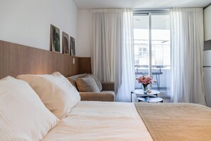 A hotel room with a bed, a sofa, a small table with a vase of flowers, and a view of the city through large windows. - Modern Studio with Pool, Gym, and Private Grill—Palermo (Buenos Aires)