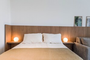 A hotel room with a wooden headboard, a bed with white bedding, two bedside lamps, and a gray armchair. - Modern Studio with Pool, Gym, and Private Grill—Palermo (Buenos Aires)