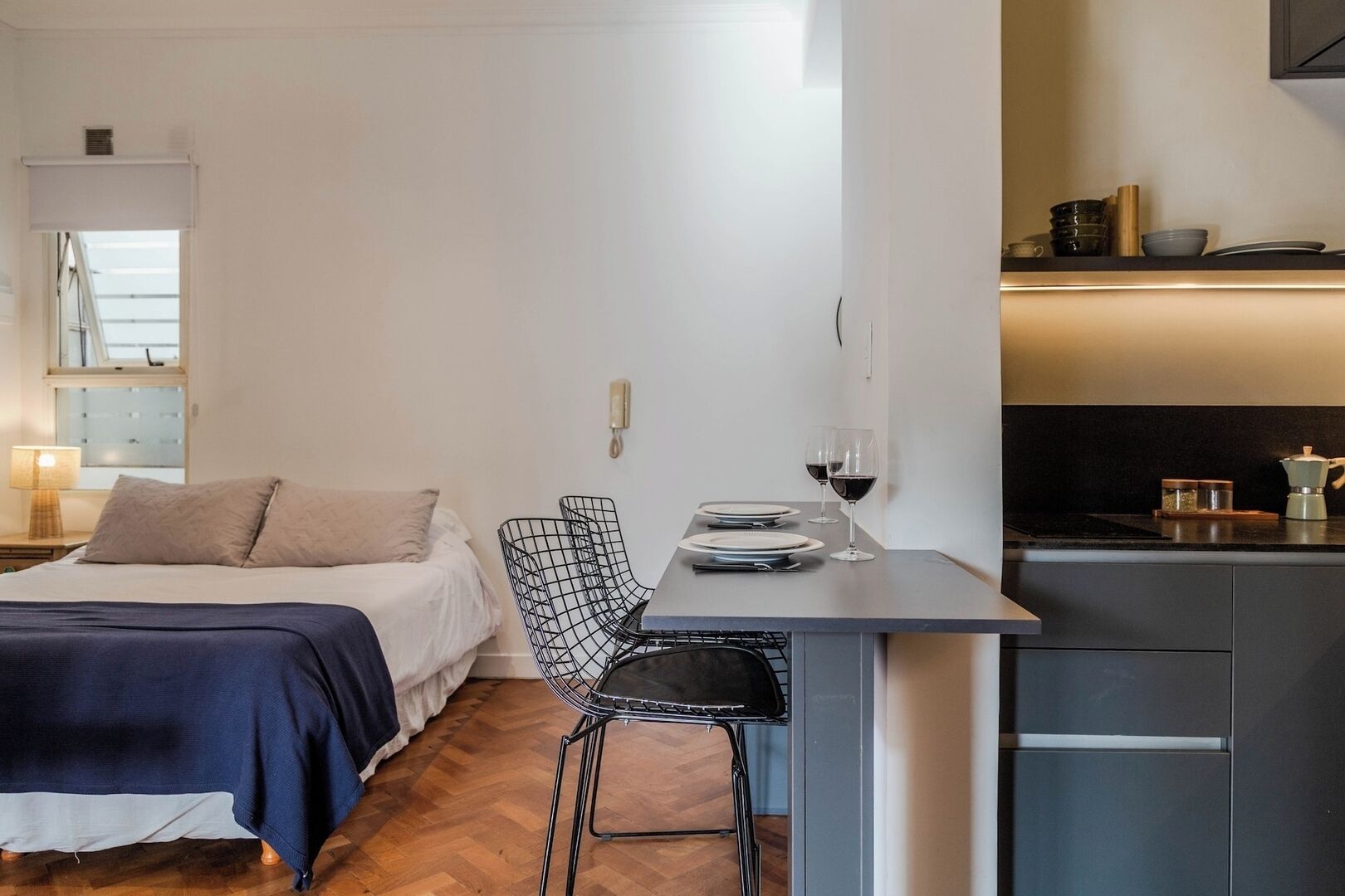 A compact hotel room with a bed, a small dining table with two chairs, a kitchen area, and a window with blinds.