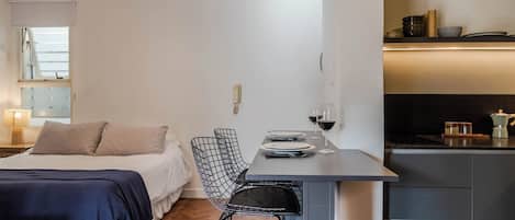 A compact hotel room with a bed, a small dining table with two chairs, a kitchen area, and a window with blinds.