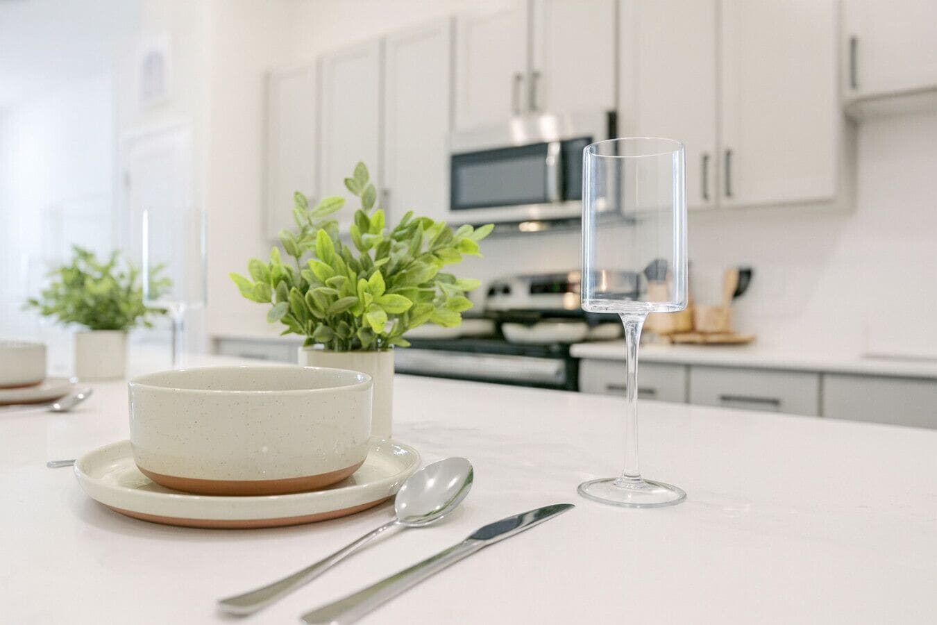 A modern kitchen with white cabinets, a microwave, and a dining table with a bowl, spoon, and glass.