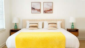 Una cama bien hecha con un edredón amarillo, dos mesitas de noche con lámparas, y dos cuadros en la pared.