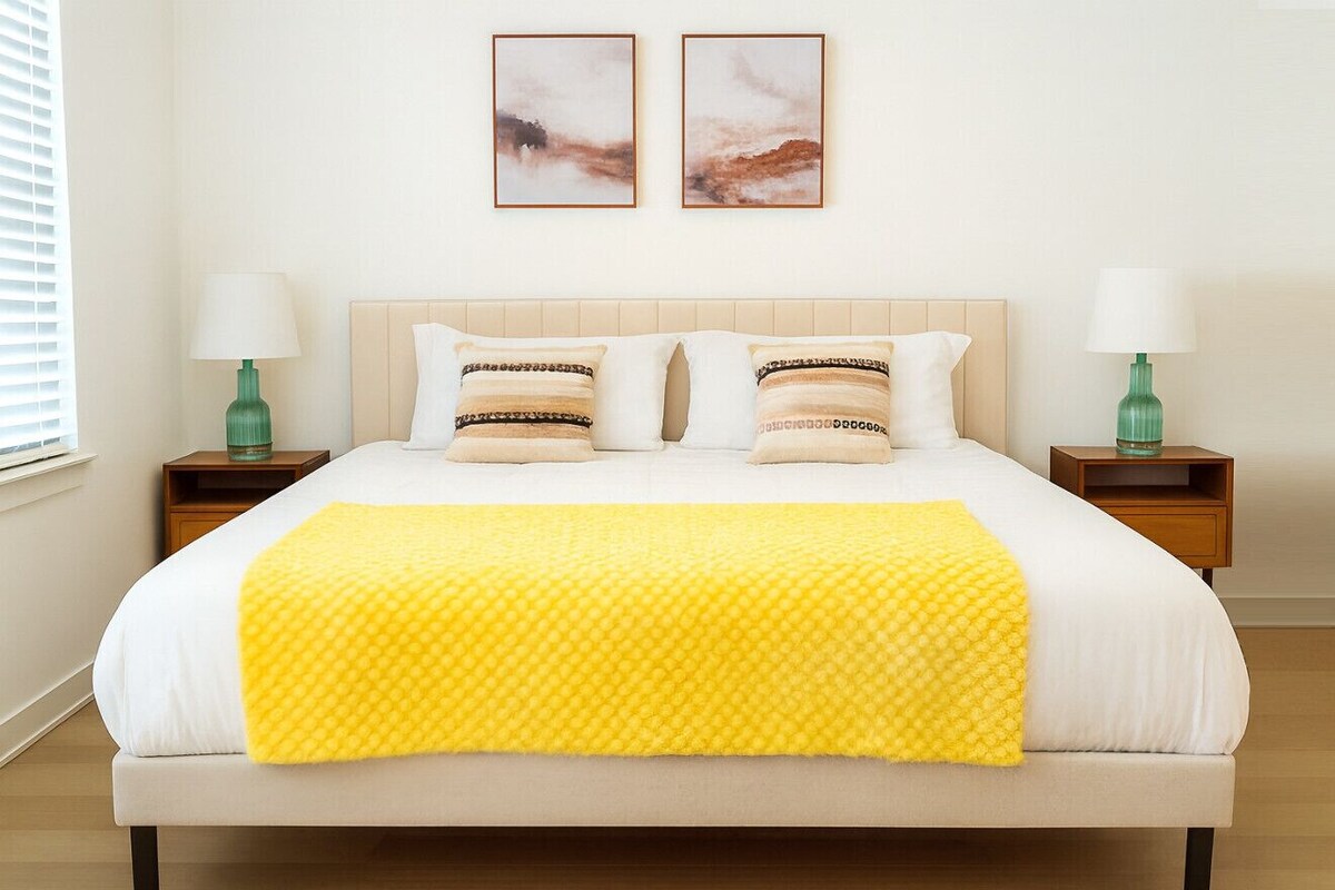 Een net opgemaakt bed met een gele deken, twee nachtkastjes met lampen en twee ingelijste foto's aan de muur.