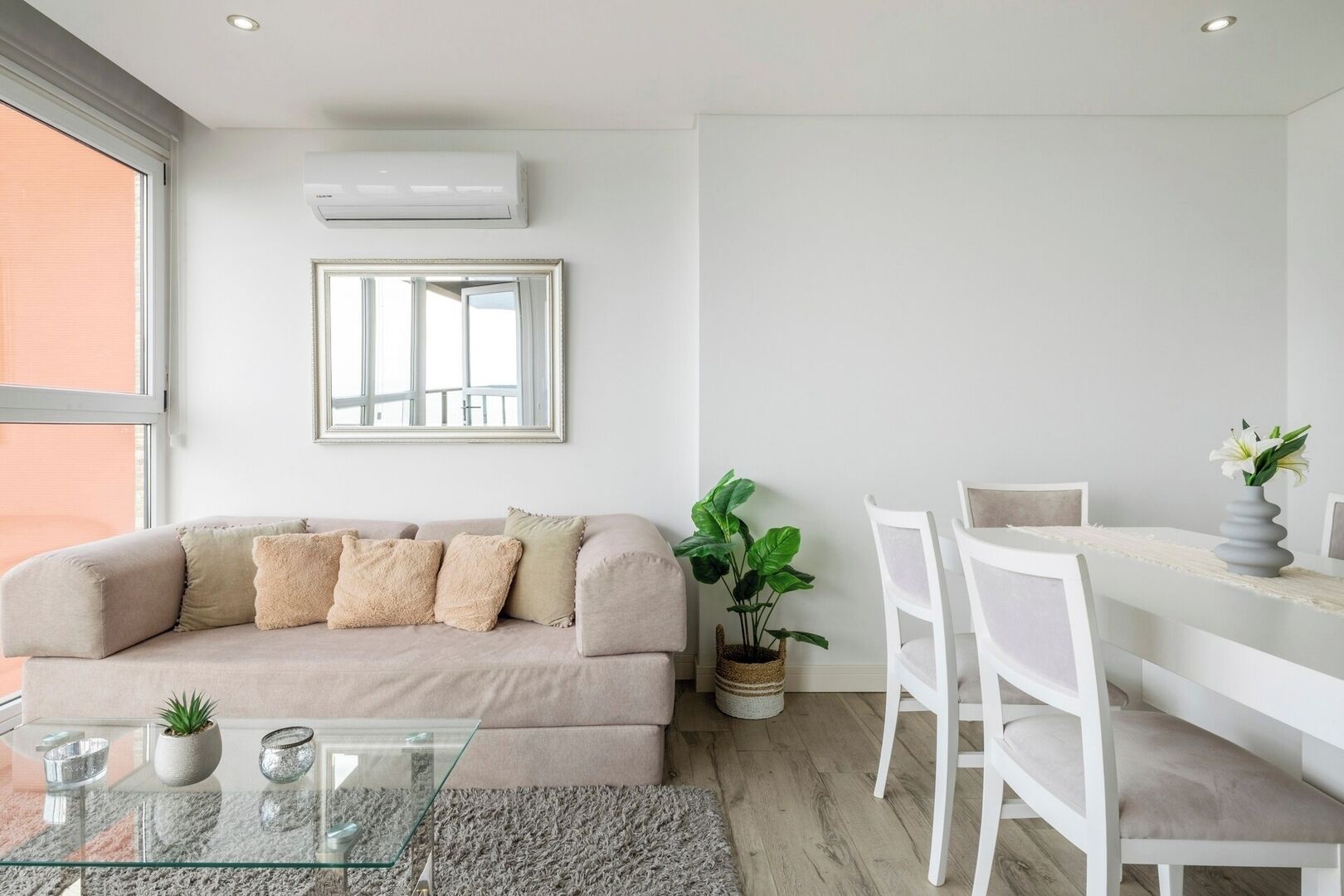 Uma sala de estar moderna com um sofá bege, uma mesa de centro de vidro, uma mesa de jantar com cadeiras brancas, um vaso de planta e um ar-condicionado fixado na parede.