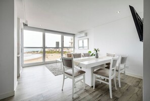 A modern dining area with a white table and chairs, large windows with a beach view, and a flat-screen TV mounted on the wall. - Bright apartment just steps from the ocean, with a pool and gym (Mar del Plata)