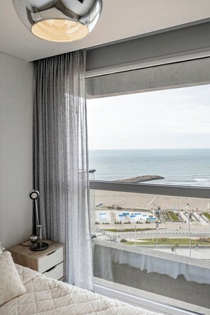 A bedroom with a large window overlooking a beach and a parking area. - Bright apartment just steps from the ocean, with a pool and gym (Mar del Plata)