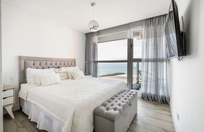 Un dormitorio con una cama grande, un banco, una mesita de noche, un televisor y vistas al mar.