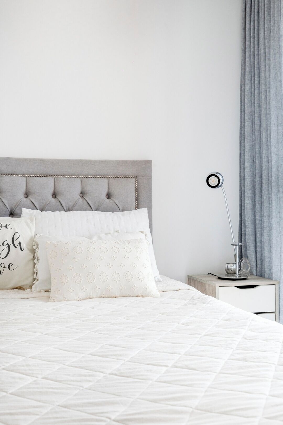 Una cama bien hecha con un cabecero capitonado, una mesita de noche con lámpara y una cortina gris.