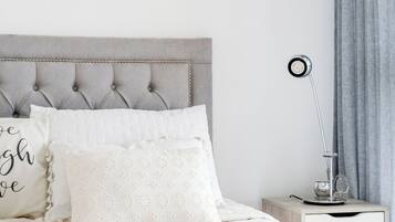 Una cama bien hecha con un cabecero capitonado, una mesita de noche con lámpara y una cortina gris.