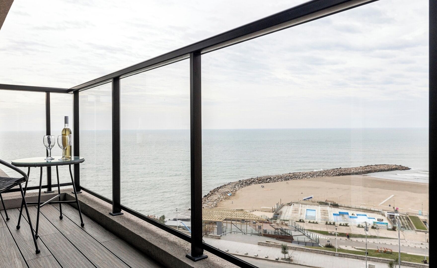 A balcony with a view of the sea, a table with a bottle and glasses, and a chair.