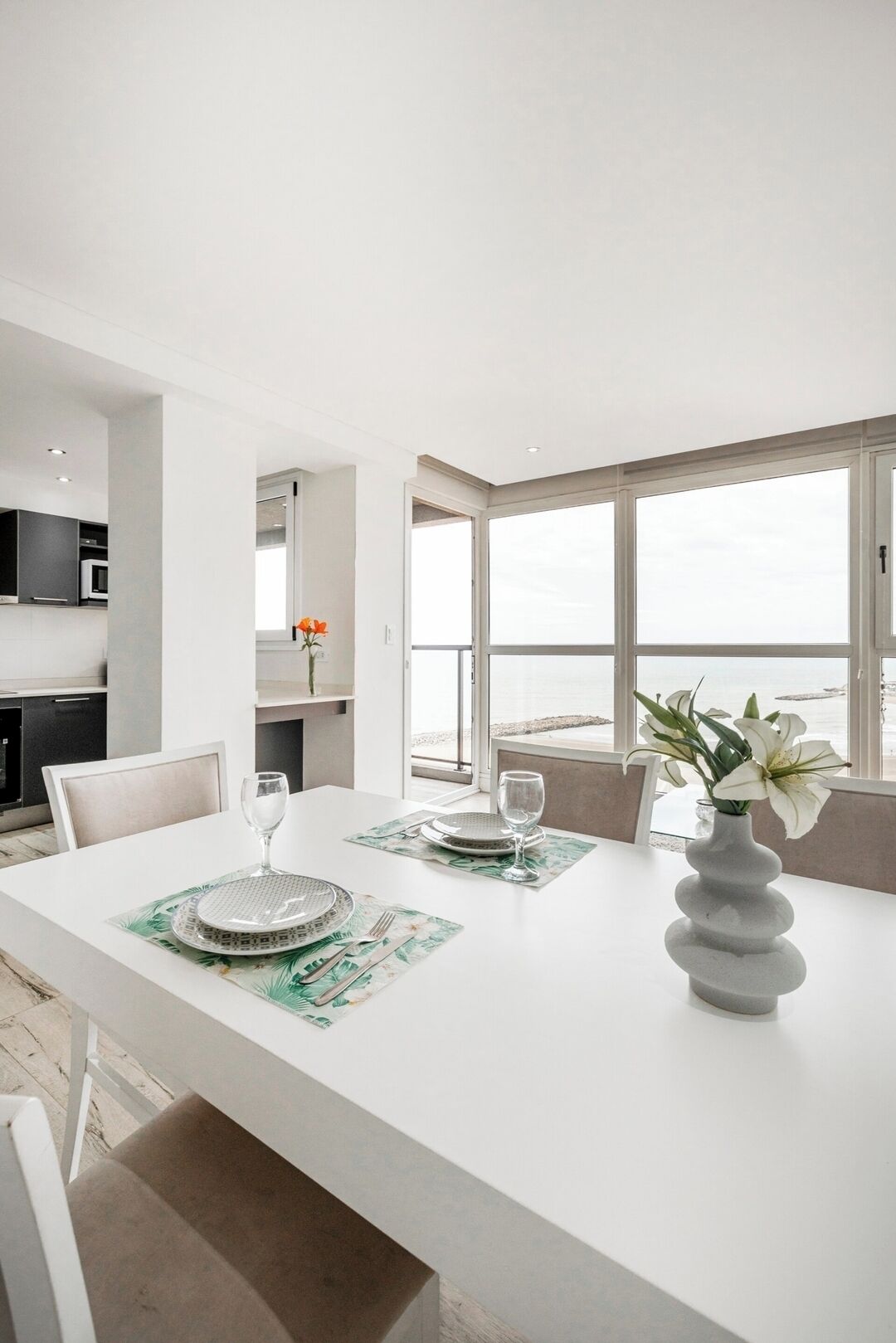 Un comedor moderno con una mesa blanca para dos, un jarrón con flores y vistas al mar a través de amplias ventanas.