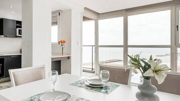 Un comedor moderno con una mesa blanca para dos, un jarrón con flores y vistas al mar a través de amplias ventanas.