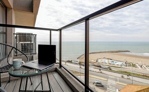Un balcon avec une table en verre, un ordinateur portable et une chaise, donnant sur une plage et une route où l’on aperçoit une voiture et un vélo.