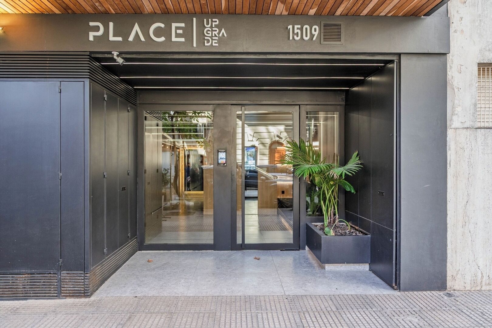 Ein moderner Gebäudeeingang mit Glastür, einem Schild mit der Aufschrift 'PLACE UPGRADE 1509' und einem Blumentopf davor.