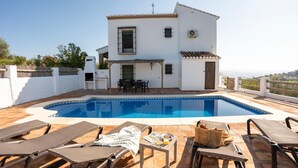 Un área junto a la piscina con sillones de descanso, una mesa con una cesta y un edificio con tejado de baldosas.