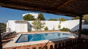 Una piscina con cubierta de madera y sillas de descanso, rodeada por un muro blanco y vegetación.