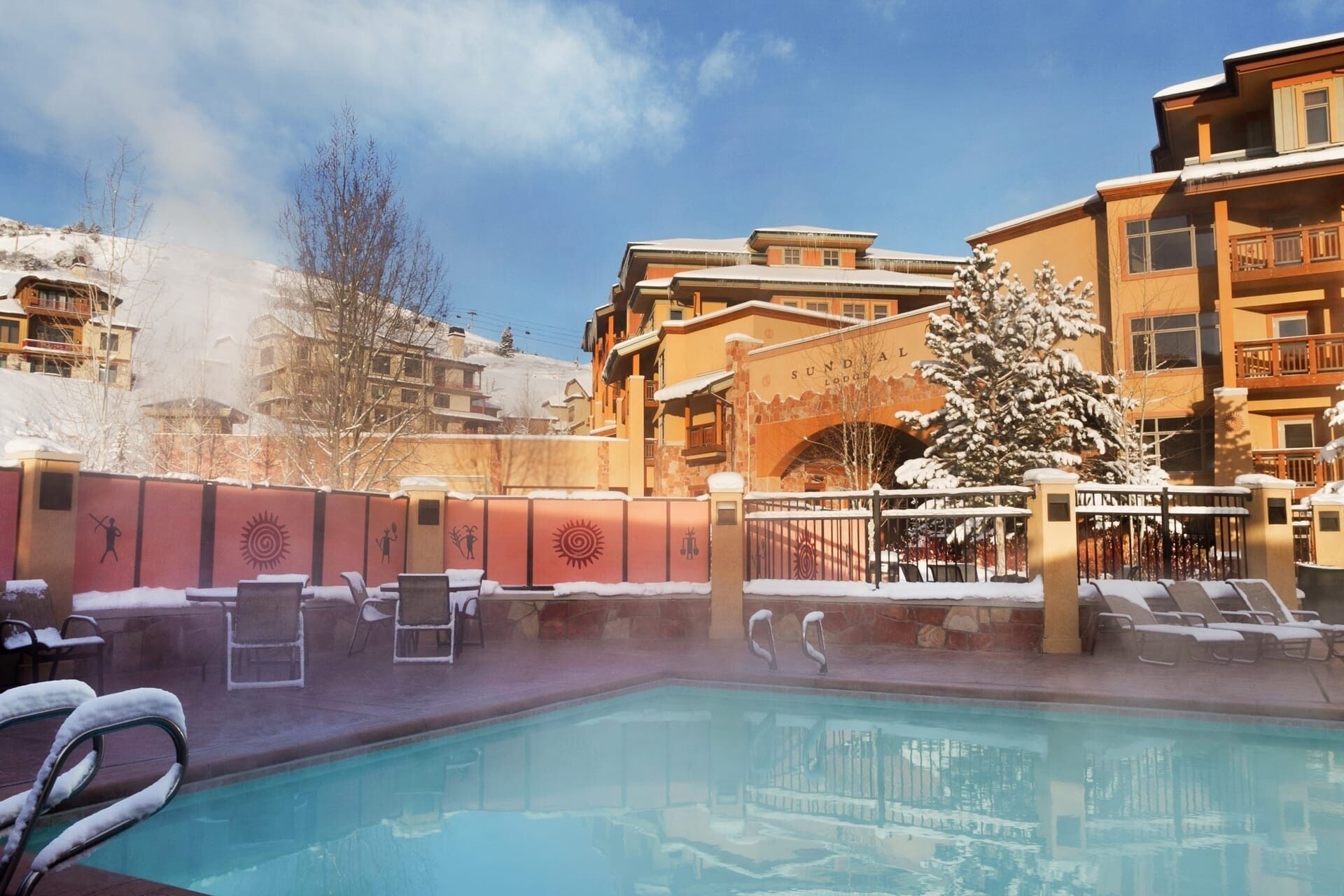 Ett poolområde med solstolar och en bubbelpool, omgivet av byggnader med en skidort som bakgrund.