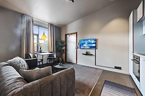 A modern living room with a sofa, a dining area with a table and chairs, a flat-screen TV mounted on the wall, and a kitchen area with built-in appliances. - Grettisborg Apartments (Reykjavik)