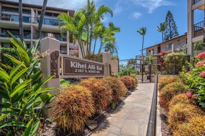 A hotel entrance with a sign, a pathway lined with plants, and a multi-story building in the background. - Kihei Alii Kai B-302 - Ocean Views, Stunning Renovation, King Size Bed, Split AC (Kihei)