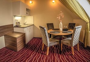 A compact hotel room with a kitchenette, a dining table with four chairs, and a red patterned carpet.