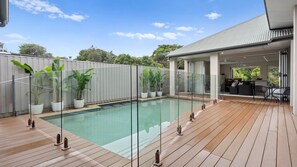 A swimming pool with a wooden deck, surrounded by potted plants, and a covered outdoor area with seating. - Collins by the Sea - Beachfront (Casuarina)