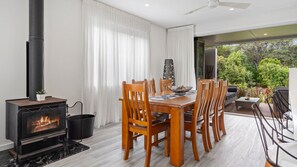 A dining area with a wood-burning stove, wooden chairs, a dining table, and a view of the outdoors. - Collins by the Sea - Beachfront (Casuarina)
