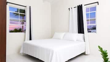 Une chambre à coucher avec un lit, des rideaux blancs et des fenêtres donnant sur des palmiers et un coucher de soleil.