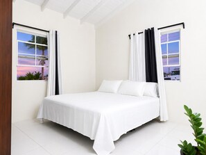 Una camera da letto con un letto, tende bianche e finestre che si affacciano su palme e un tramonto.