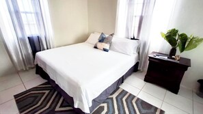 A bedroom with a bed, pillows, a nightstand, a plant, and a window with curtains. - Peaceful 2-bedroom house with AC in Christ Church Parish (Christ Church Nichola Town Parish)