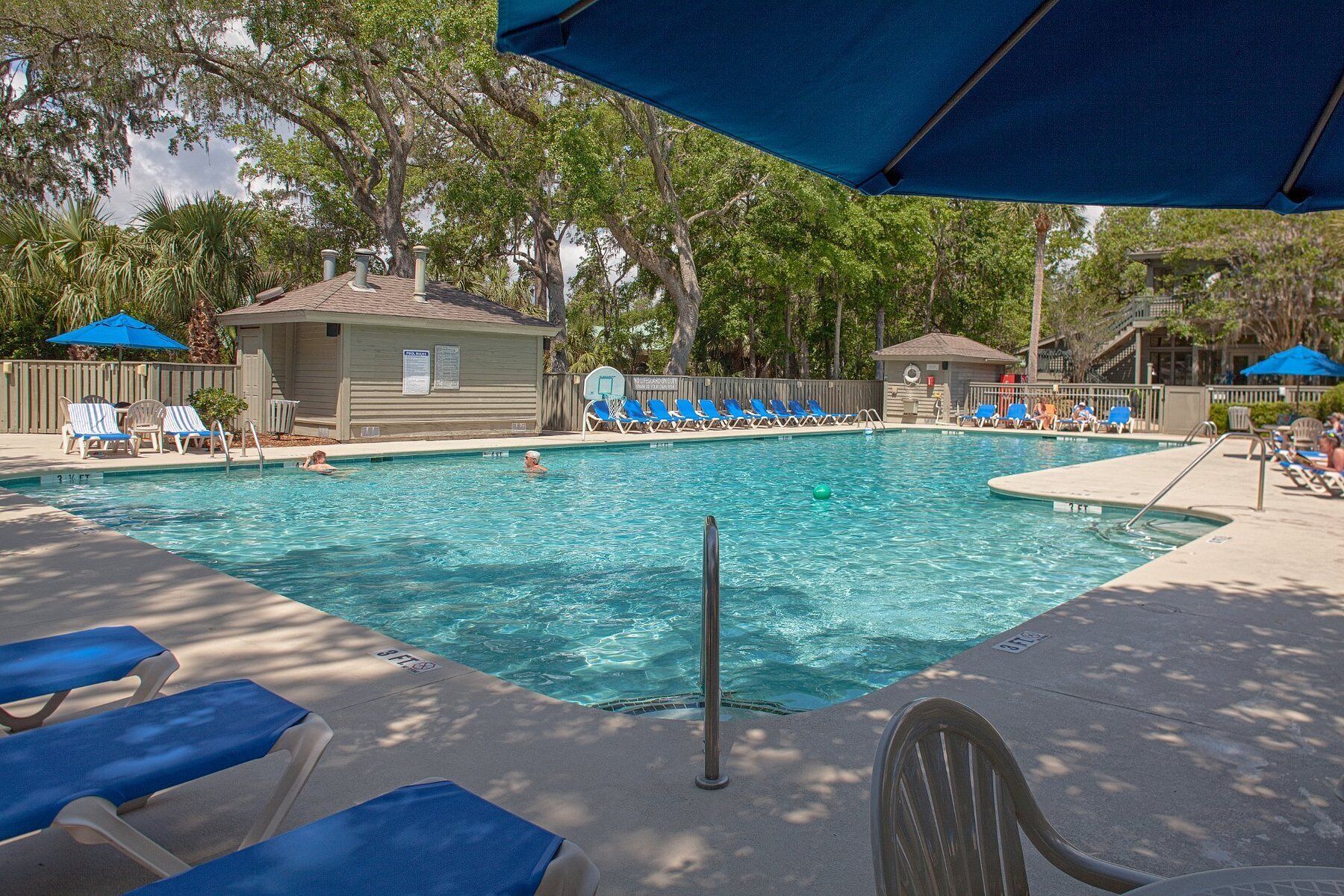 A swimming pool with lounge chairs and umbrellas, surrounded by trees and a building.