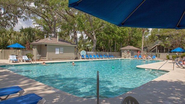 Une piscine entourée d’arbres et d’un bâtiment, avec des chaises longues et des parasols à proximité.