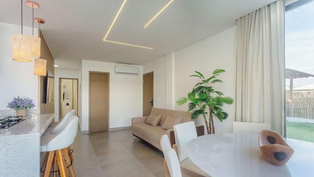 A modern living room with a sofa, a dining table, and chairs. There is a potted plant and a large window with curtains. - Apt With Pool View, Mins From the sea Vll0001 (Maraú)