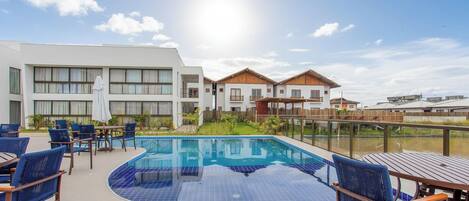 Una piscina con mosaicos azules, rodeada de sillones y mesas, con edificios al fondo.