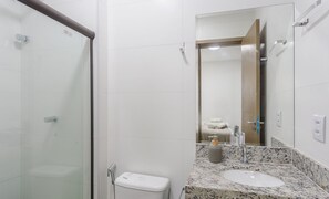 Un baño con cabina de ducha de vidrio, inodoro, lavamanos con encimera de mármol y espejo que refleja un perchero y una puerta.