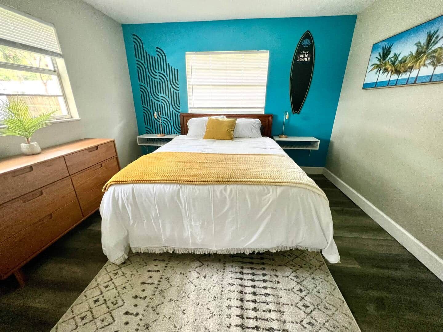 Une chambre avec un lit, une commode, une fenêtre, une planche de surf et un mur orné d’une fresque aux motifs tropicaux.